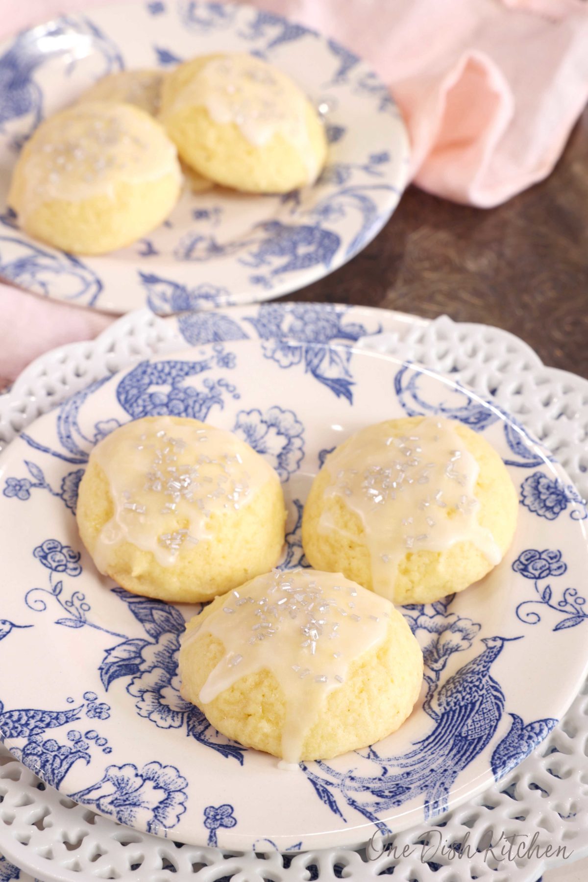 three ricotta cookies on a blue ad white plate with another plate of cookies in the background next to a pink napkin.