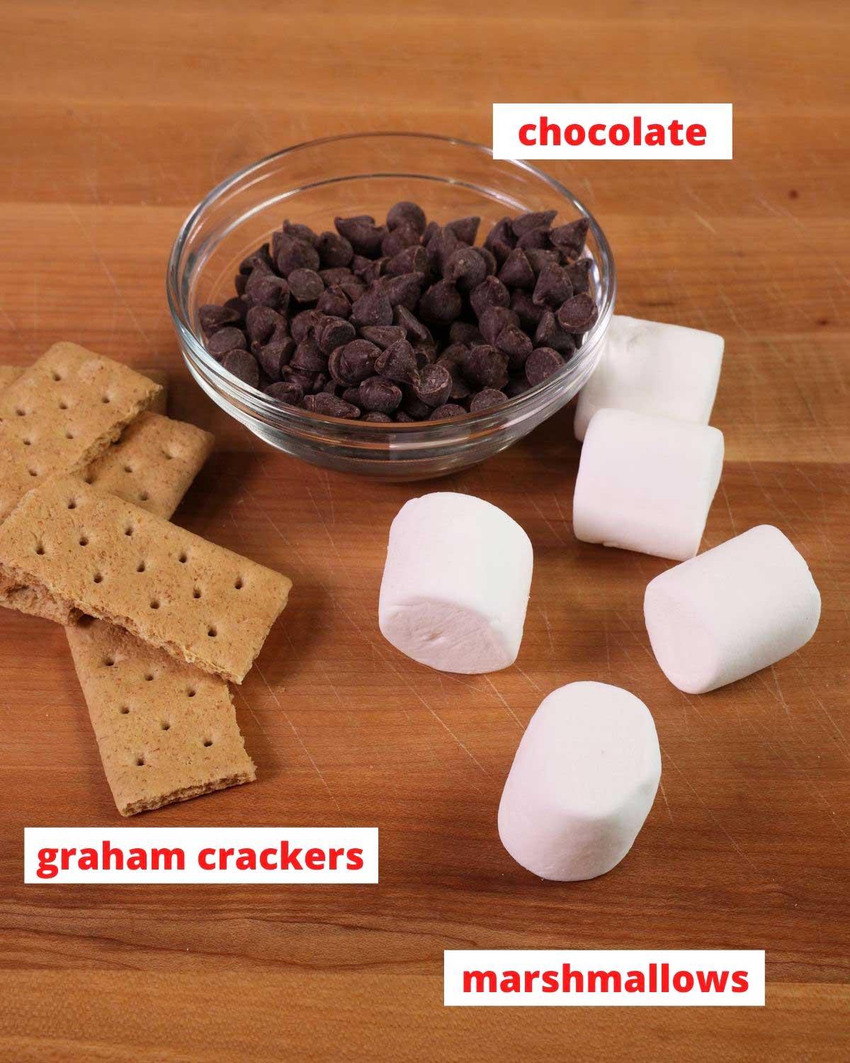 chocolate chips, graham crackers and marshmallows on a brown wooden cutting board.