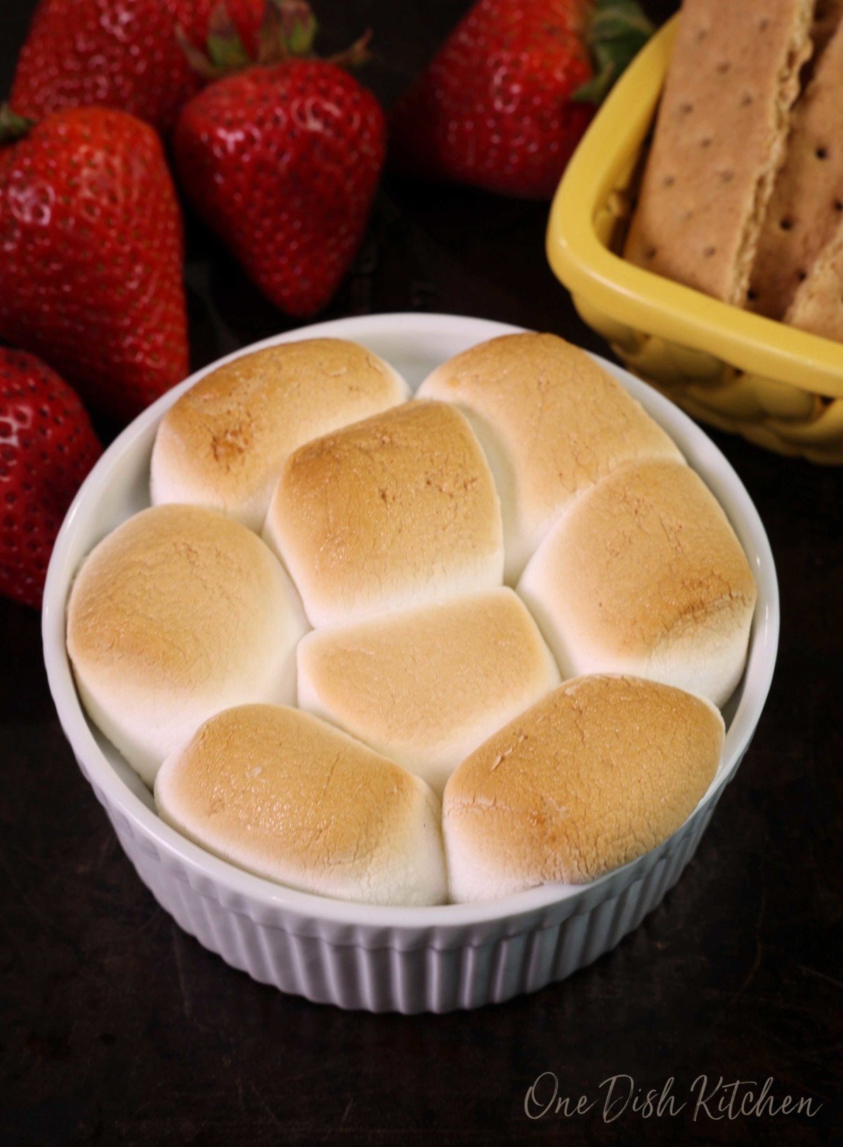 a smores dip on a black tray next to strawberries and a bowl of graham crackers.