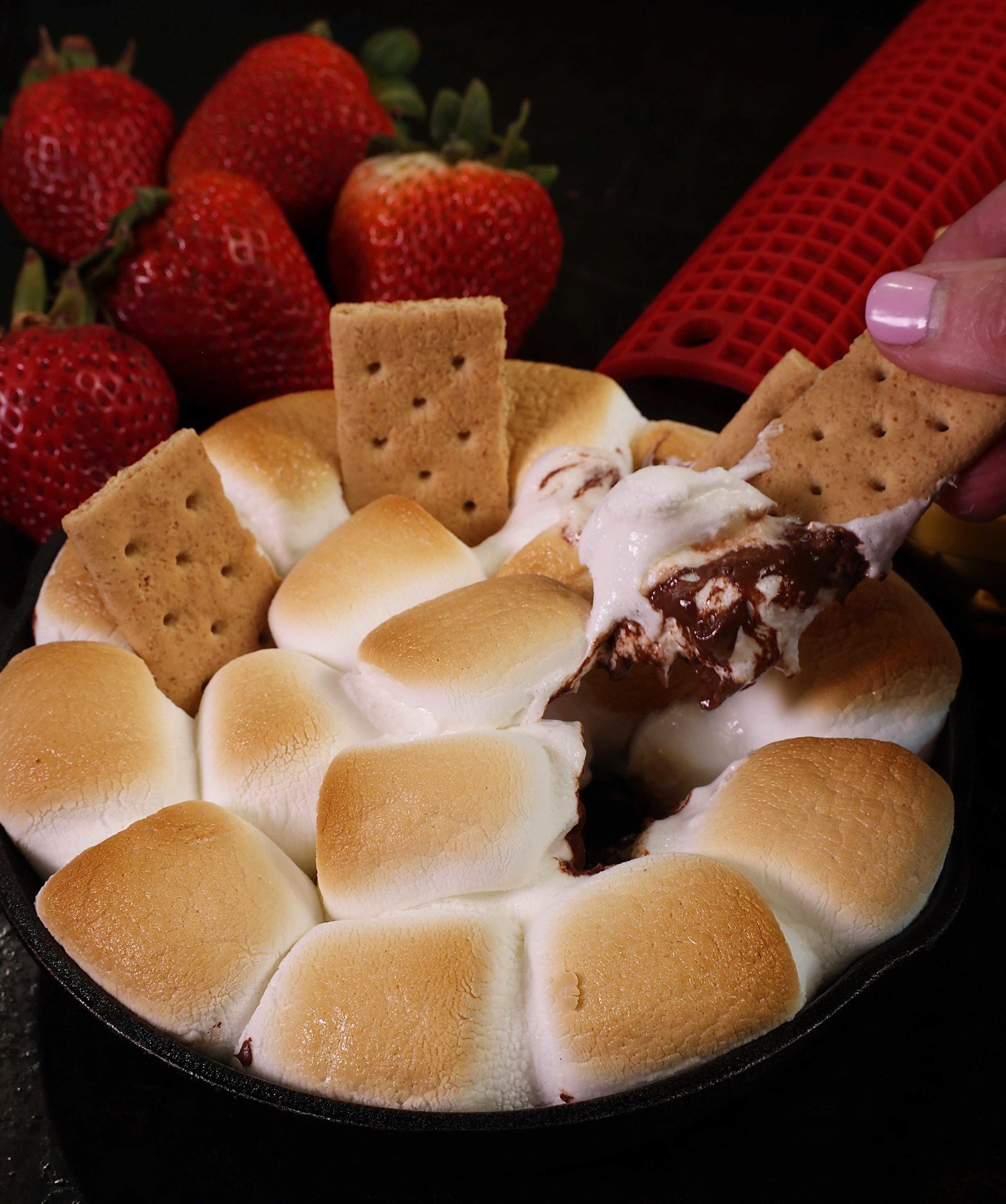 a graham cracker dipped into a skillet filled with melted chocolate and toasted marshmallows