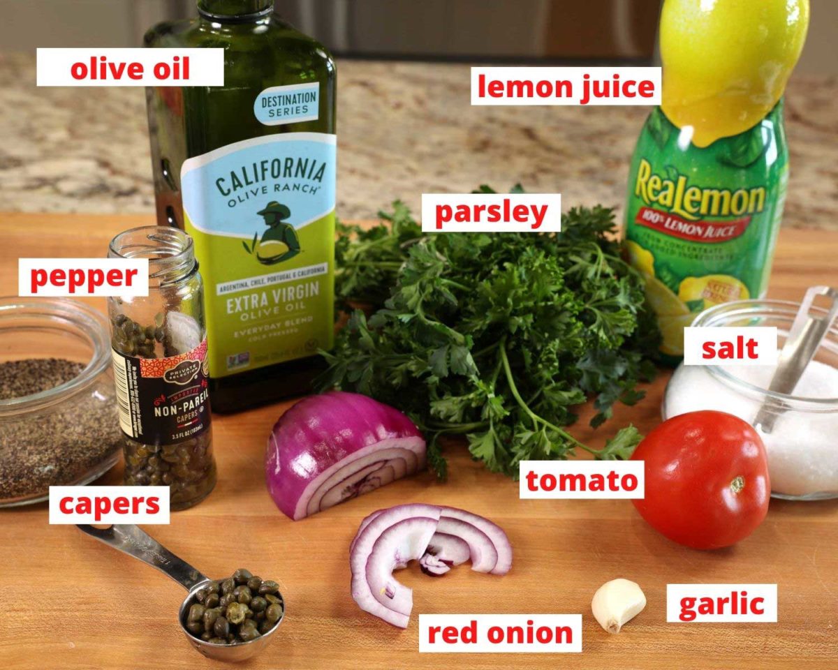 a bottle of olive oil, lemon juice, onions, tomato, parsley, and capers on a wooden cutting board.