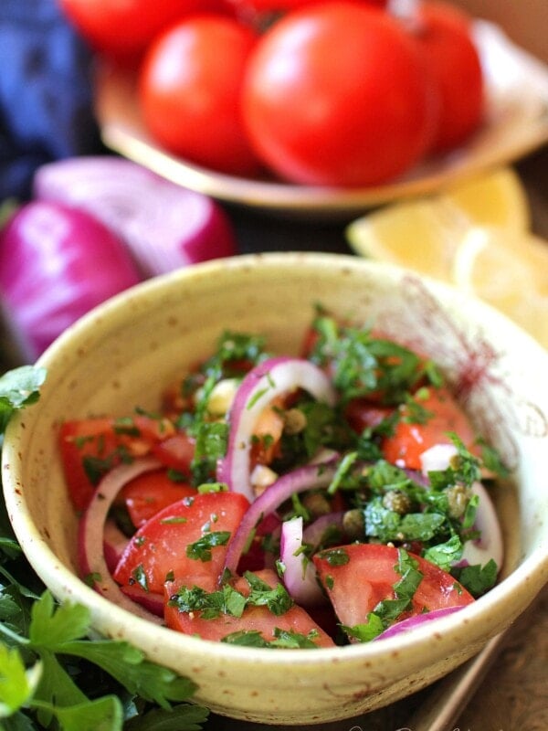 a yellow bowl filled with chopped tomatoes, sliced red onions,a nd chopped parsley next to lemon wedges and a bowl full of red tomatoes.