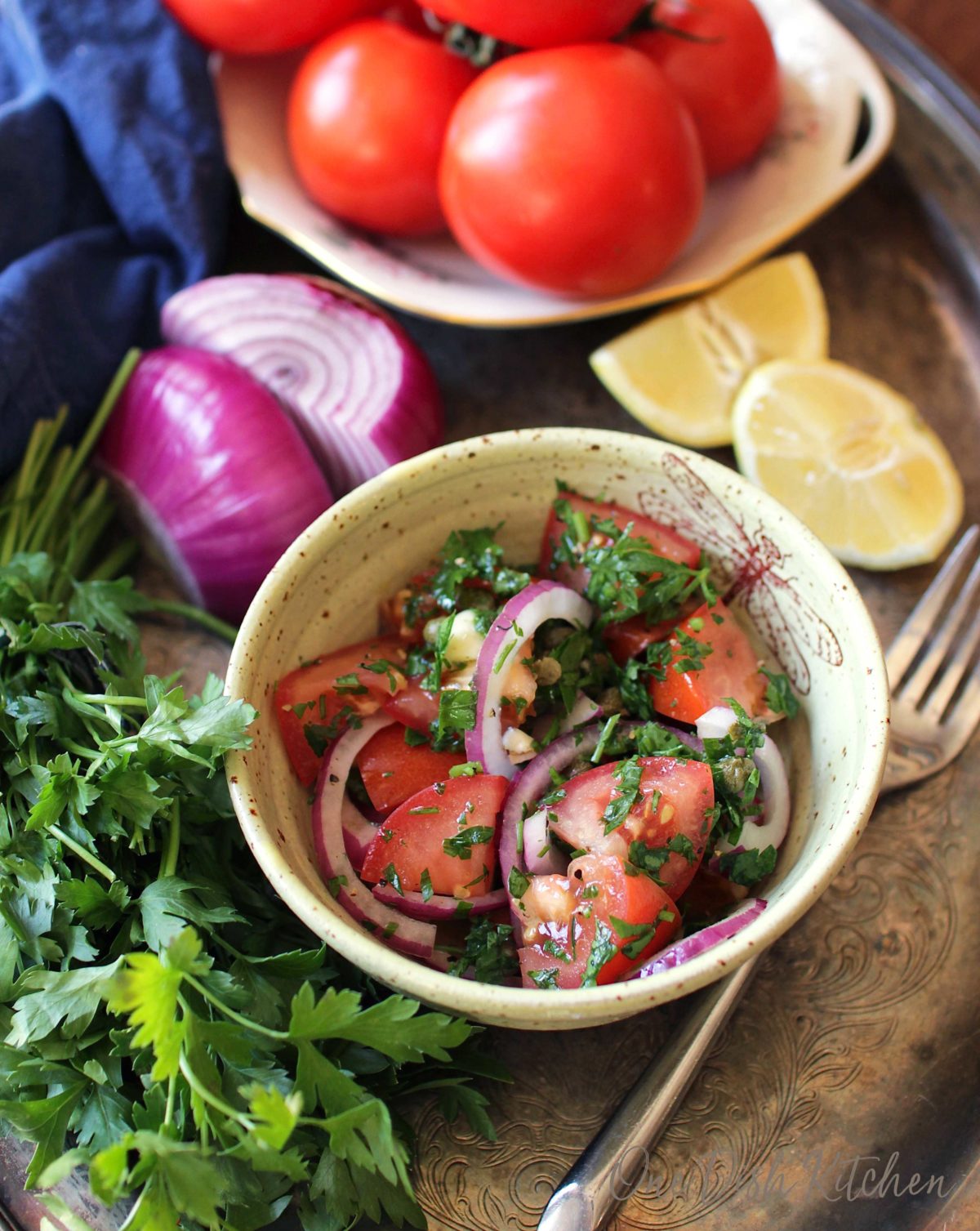 a tomato salad in a yellow bowl on a silver tray surrounded by a red onion sliced in half, a bowl of fresh tomatoes, lemon slices, and parsley.