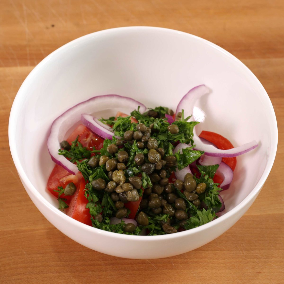 a white bowl filled with capers, tomatoes, red onion slices, and parsley on a brown wooden table.