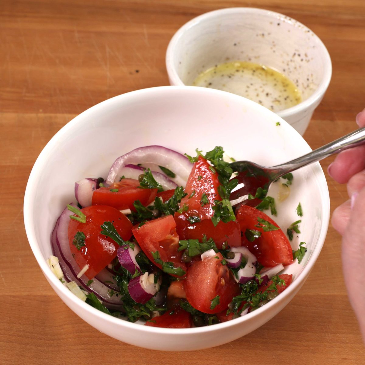 a white bowl filled with a tomato salad and a fork on the side of the bowl. In the background is an empty white bowl with a few drops of remaining dressing inside.
