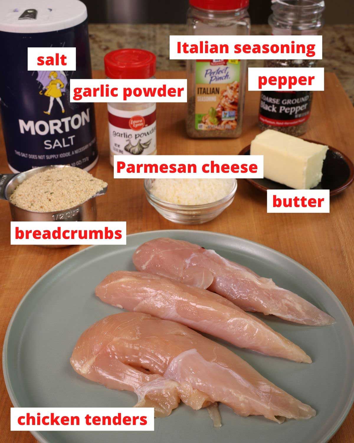 three strips of chicken on a green plate next to a bowl of breadcrumbs, parmesan cheese, butter and jars of salt, pepper, and italian seasoning