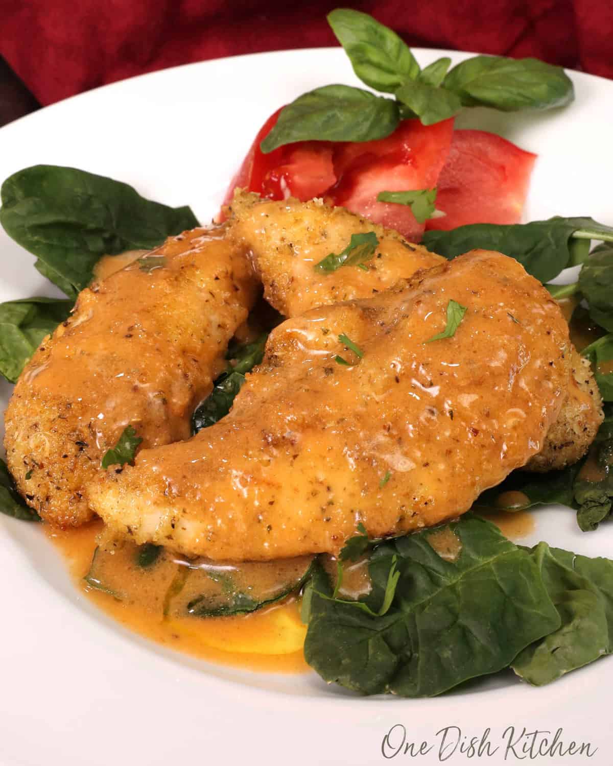 three chicken tenders topped with buffalo sauce on a bed of fresh spinach next to a sliced tomato on a white plate.