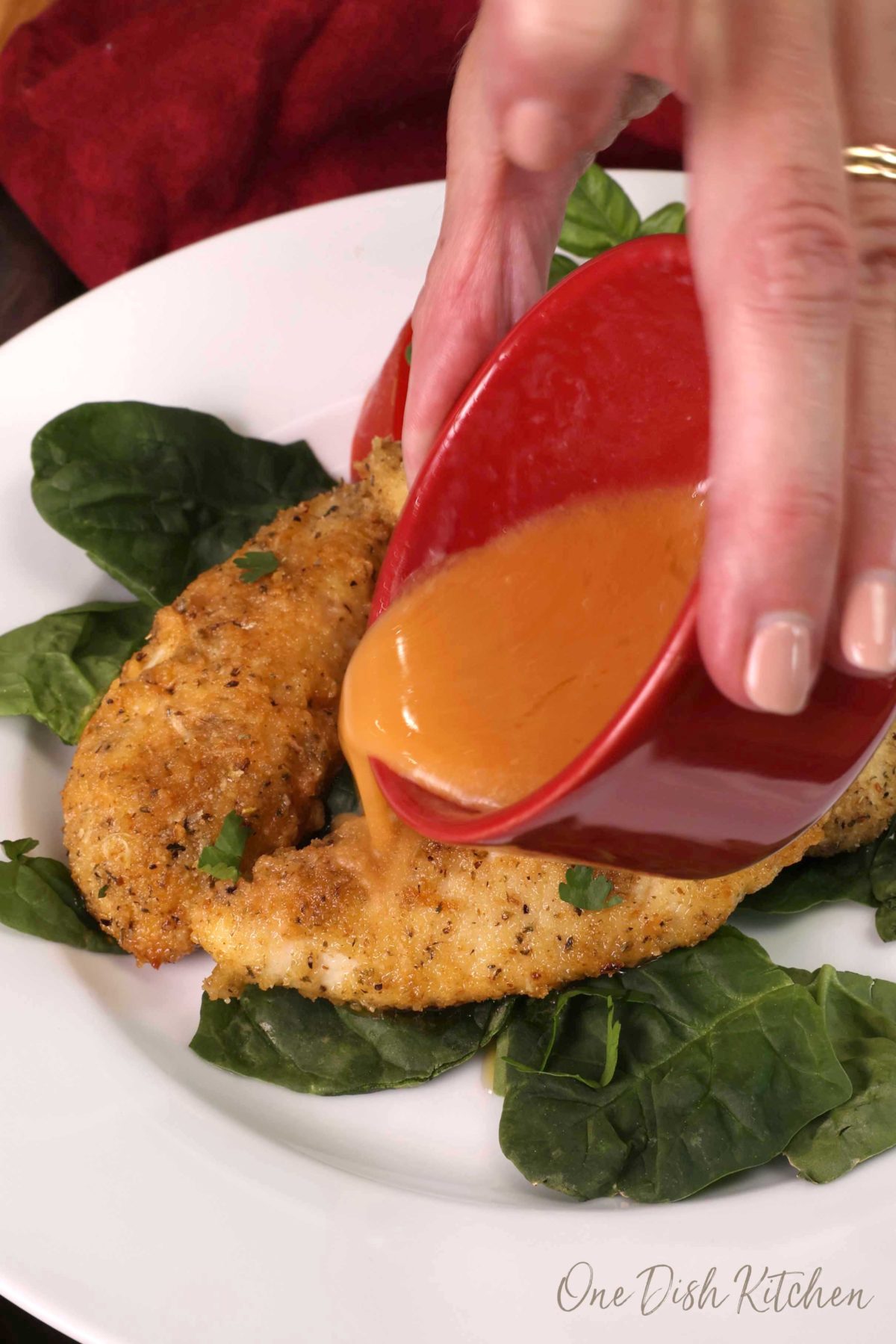 three breaded pieces of chicken on a white plate with buffalo sauce from a red bowl being poured over them.