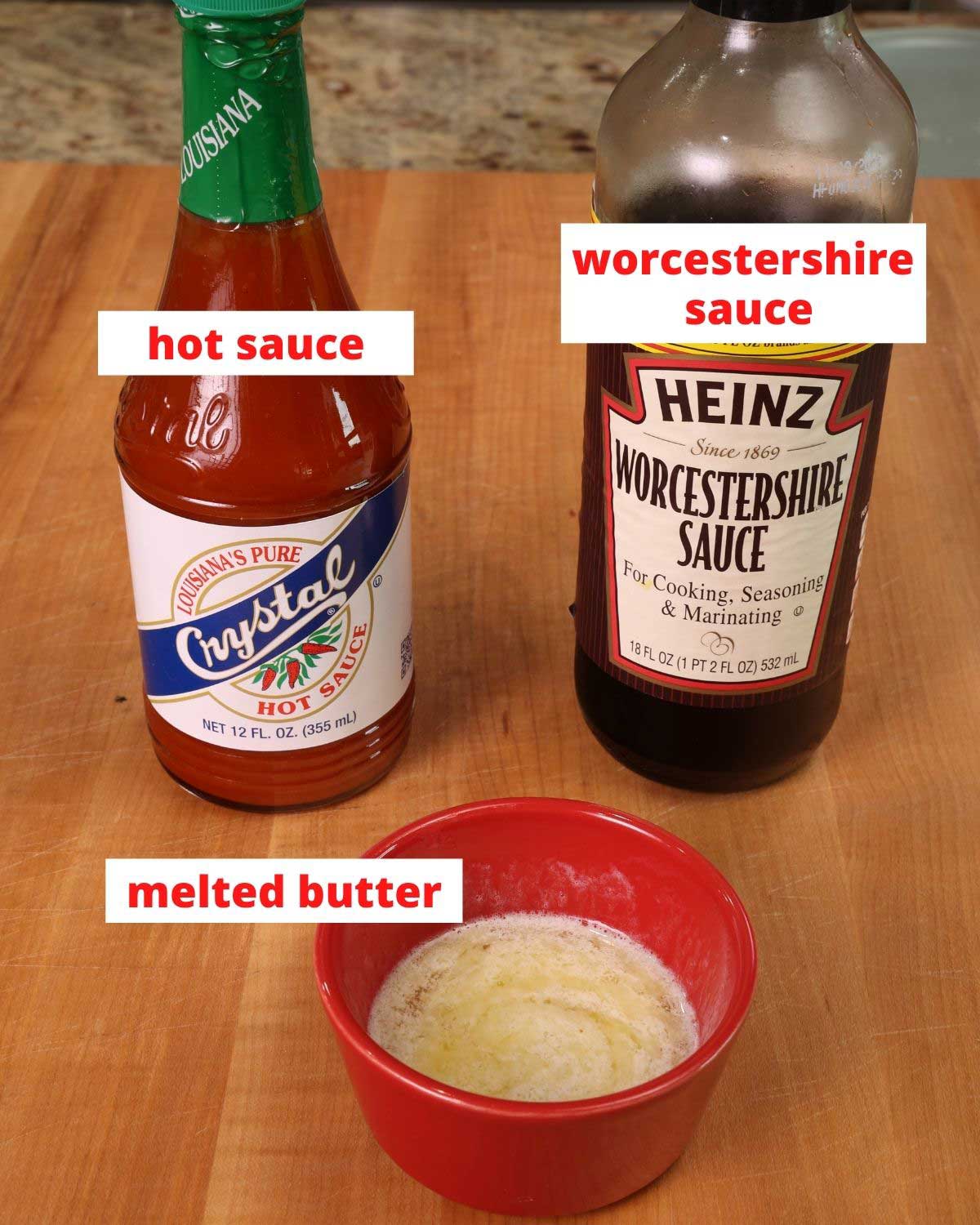 a bottle of hot sauce, worcestershire sauce and a bowl of melted butter on a brown wooden cutting board
