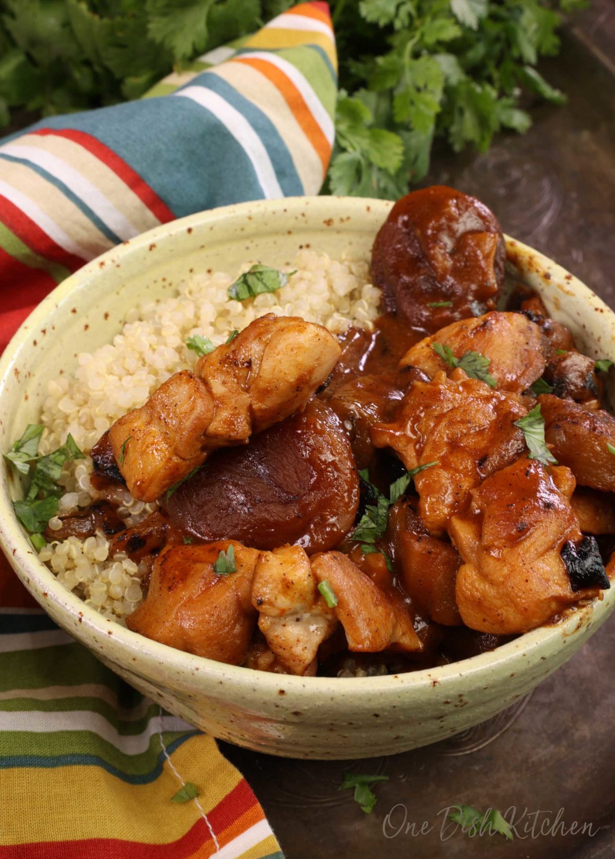 a yellow bowl filled with eggplant and chicken tagine over quinoa next to a striped red and blue napkin on a silver tray.
