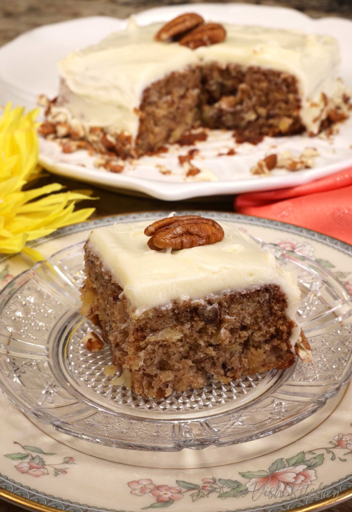 a square piece of hummingbird cake on a white plate next to a yellow flower and the remaining cake on a white plate.