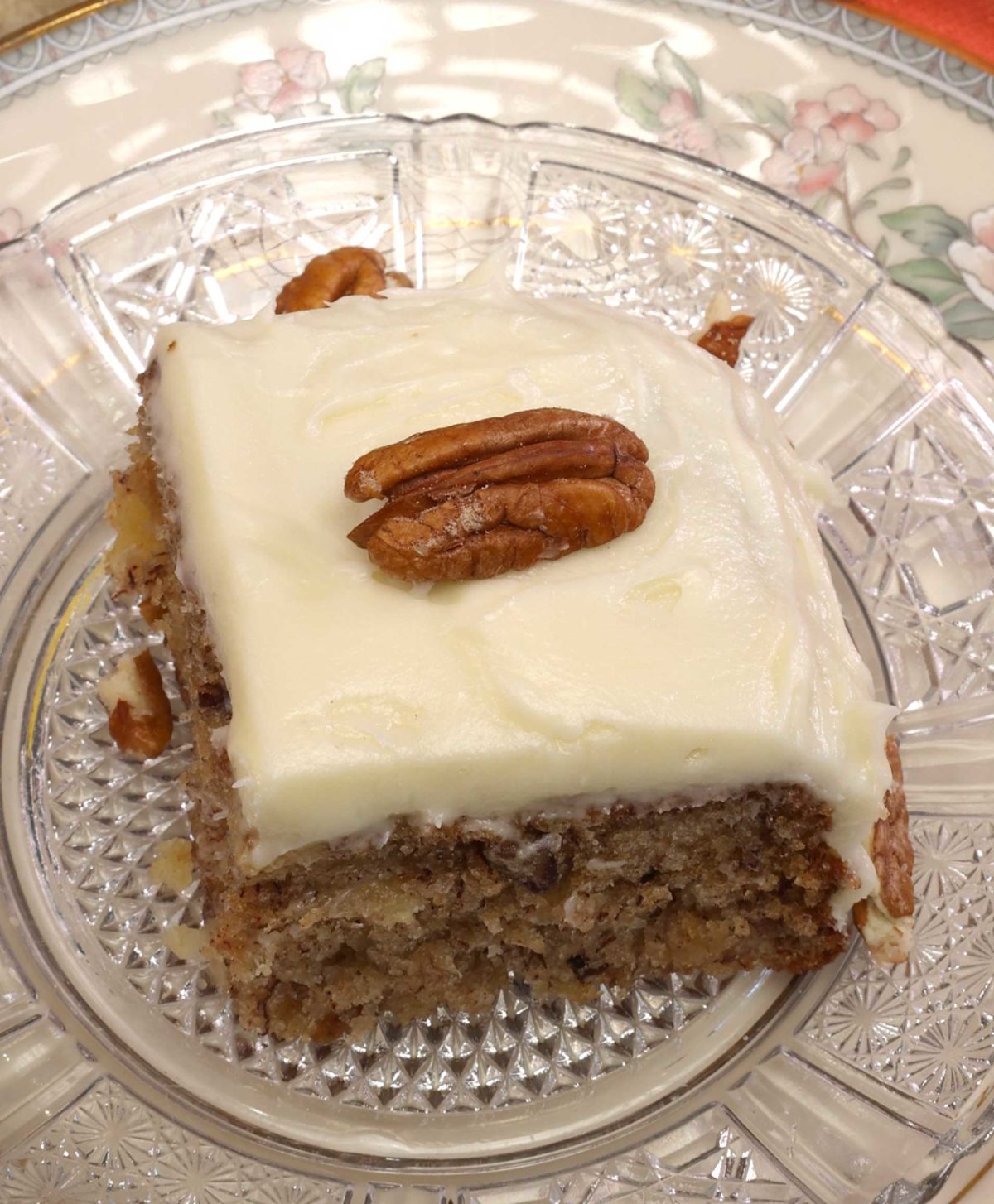 a square piece of hummingbird cake on a white plate with a pecan on the top of the cake.