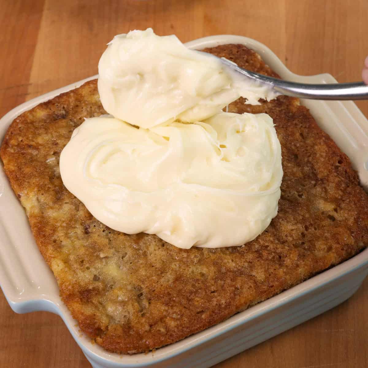 a small hummingbird cake in a square baking dish with a dollop of cream cheese frosting on top.