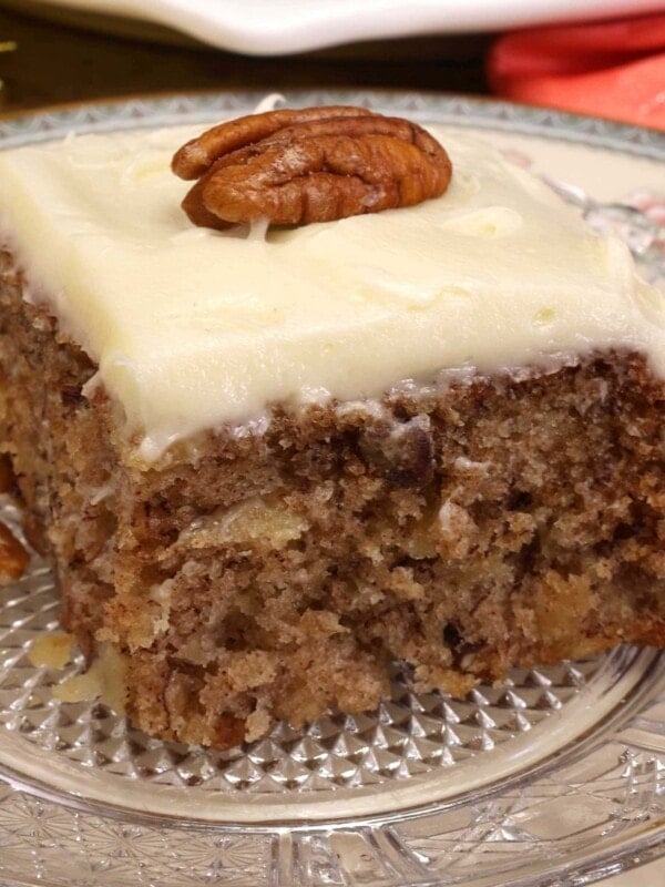 a slice of hummingbird cake topped with cream cheese frosting and a pecan on the top sitting on a clear plate