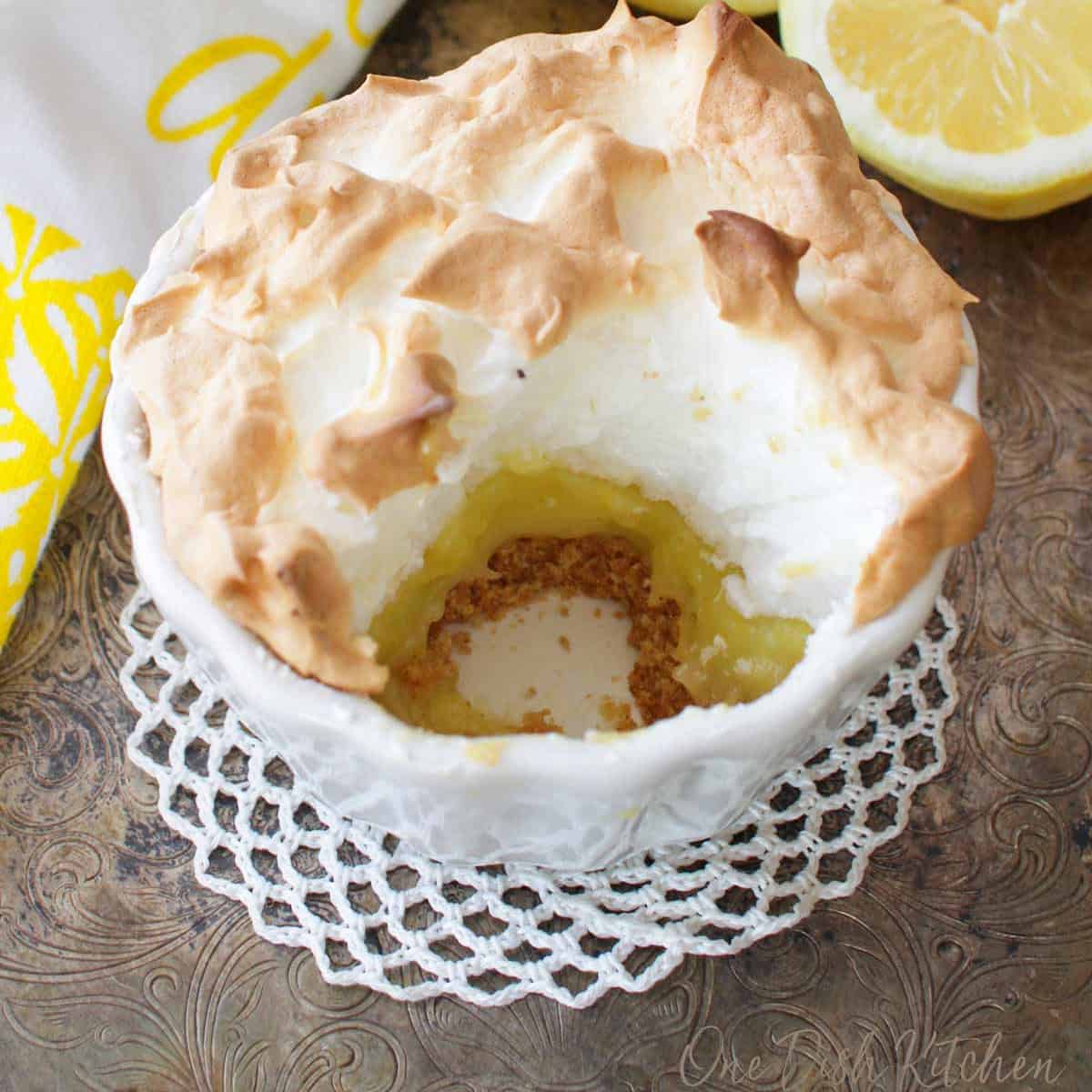 A small round lemon meringue pie with a round section removed next to sliced lemons on a silver tray.