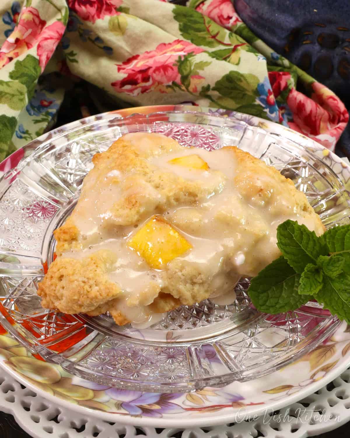 one peach scone on a floral plate next to a sprig of fresh mint and a flowered napkin in the background.