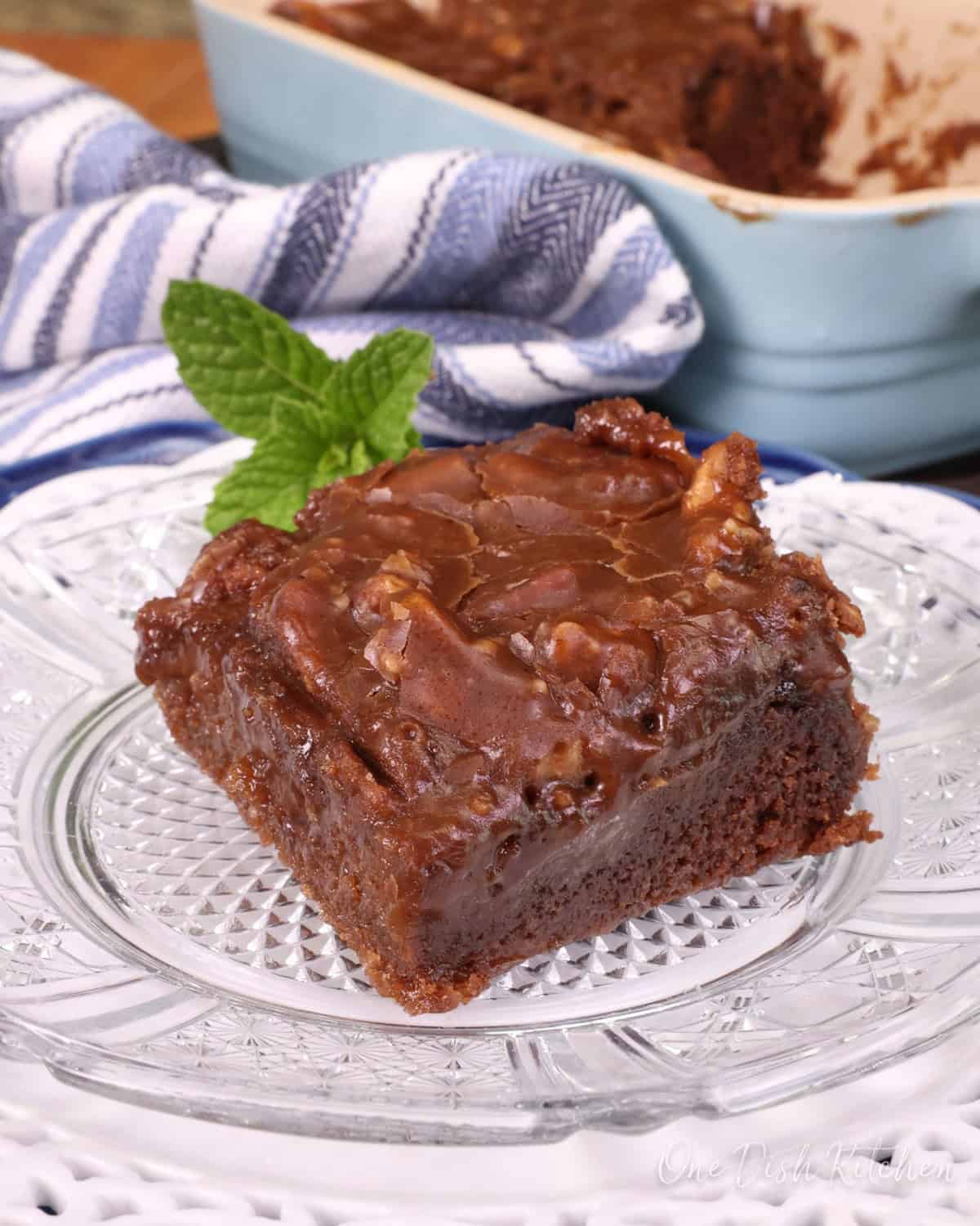 a square piece of chocolate cake on a white plate next to a larger rectangular blue baking dish with the remaining slices of cake inside.