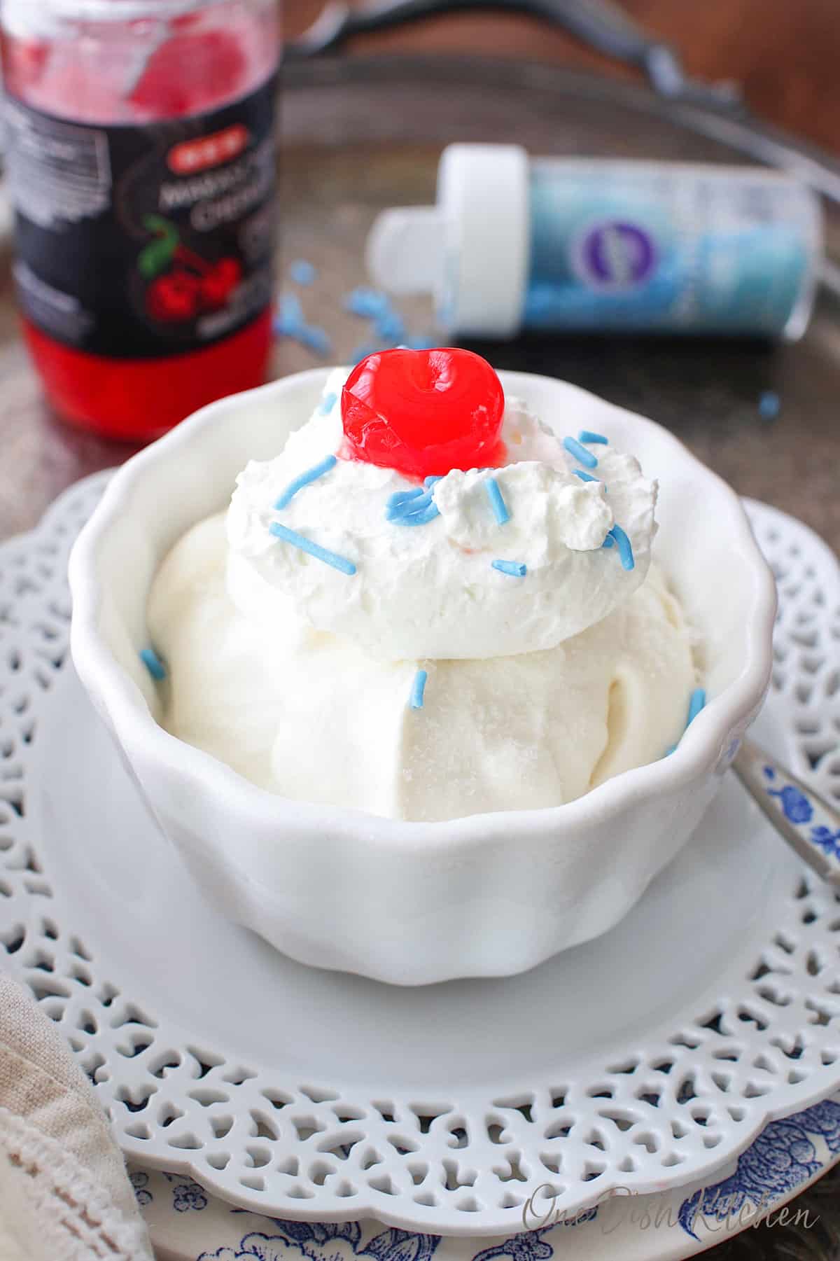 a white bowl filled with vanilla ice cream and topped with blue sprinkles and one maraschino cherry.