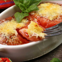 two broiled tomatoes in a white oval baking dish next to a fork and fresh basil