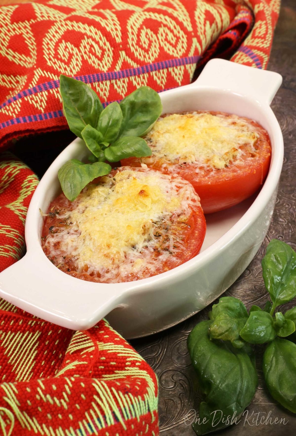 two broiled tomatoes in a white oval dish topped with melted parmesan cheese and fresh basil.