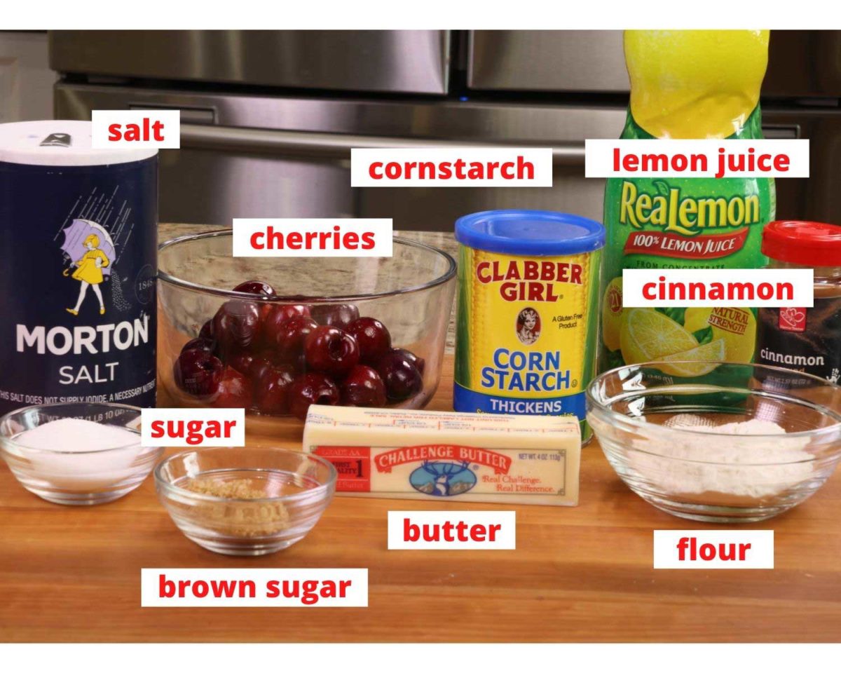 a bowl of cherries surrounded by flour, sugar, butter, cornstarch, and lemon juice on a large wooden cutting board.