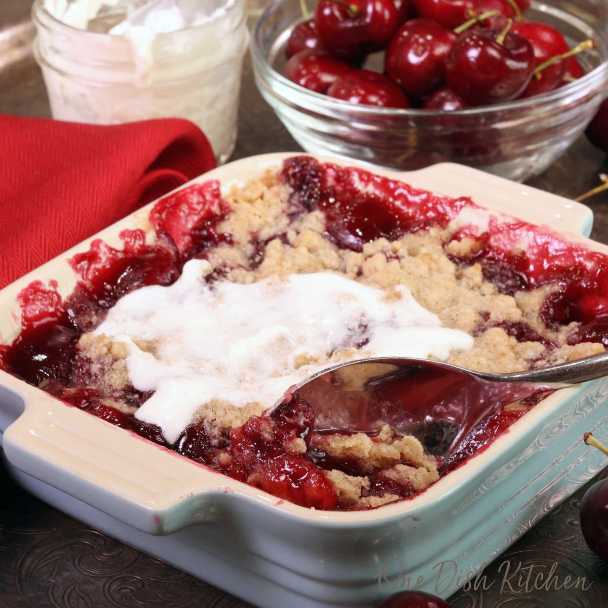 a spoon inserted in the corner of a cherry pie next to a bowl of fresh cherries and a jar of whipped cream.