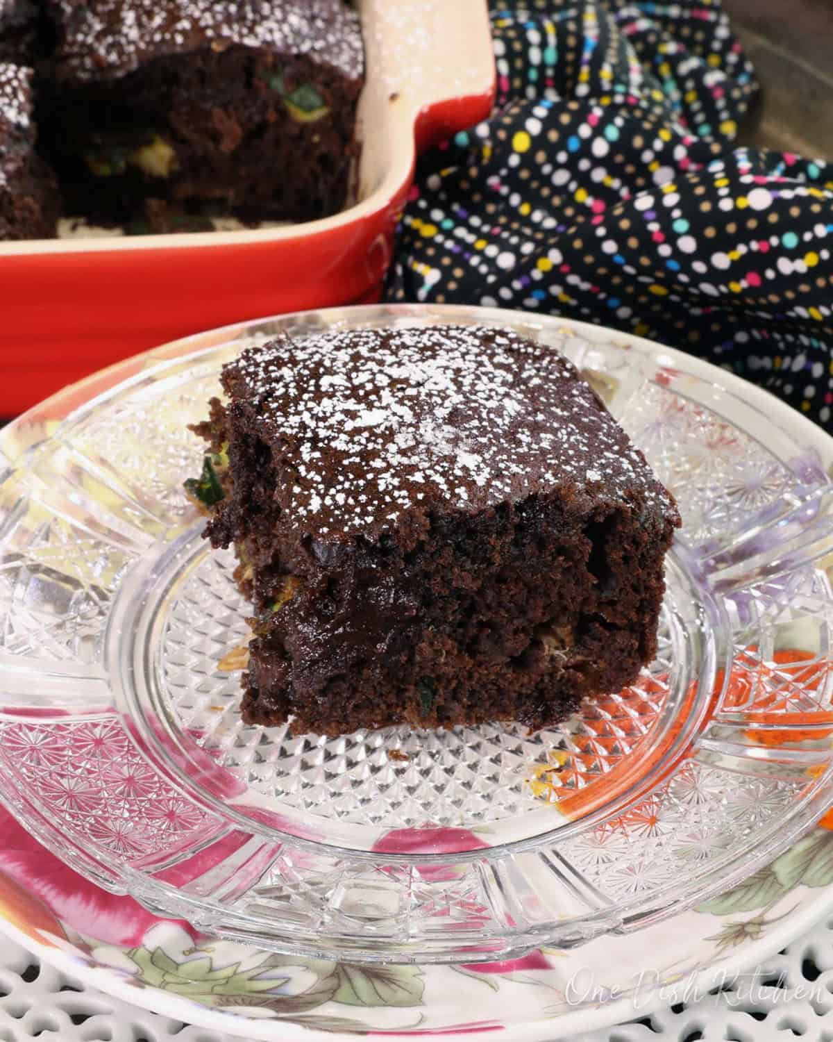 a slice of chocolate zucchini bread on a floral plate next to a red baking dish with the remaining bread inside.