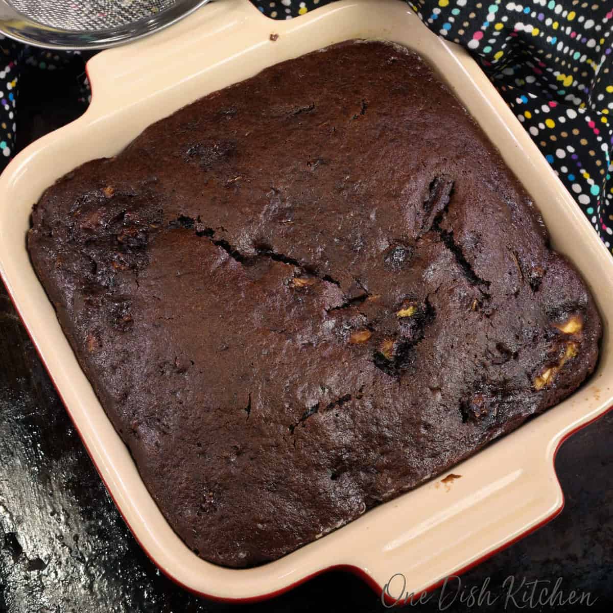 chocolate cake with zucchini in a red baking dish next to a jar of powdered sugar.