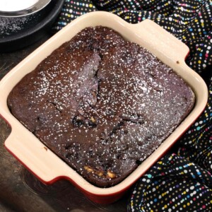 an overhead photo of a chocolate zucchini bread topped with powdered sugar in a small red baking dish next to a multi-colored dish towel