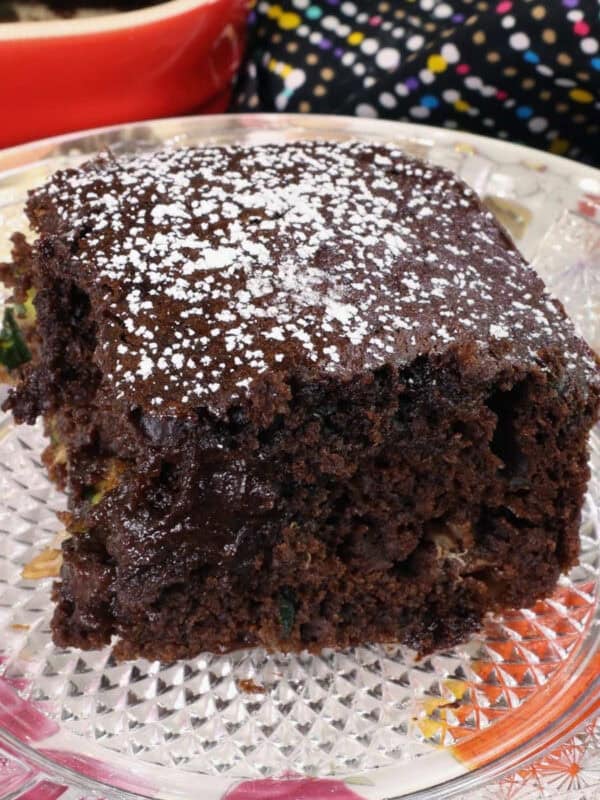 a slice of chocolate zucchini bread on a floral plate next to a red baking dish with the remaining bread inside.