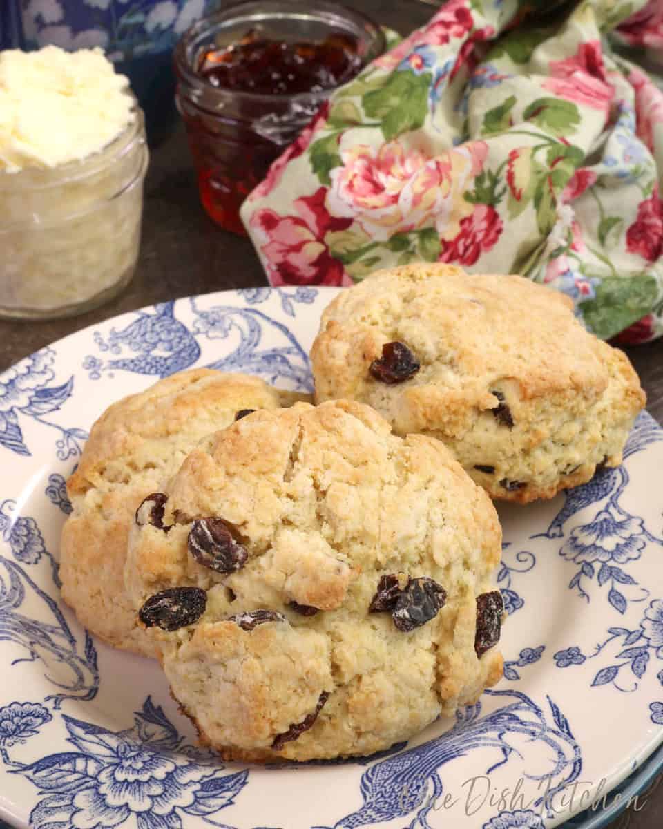 two cream scones with raisins on a blue and white plate with a jar of clotted cream in the background.