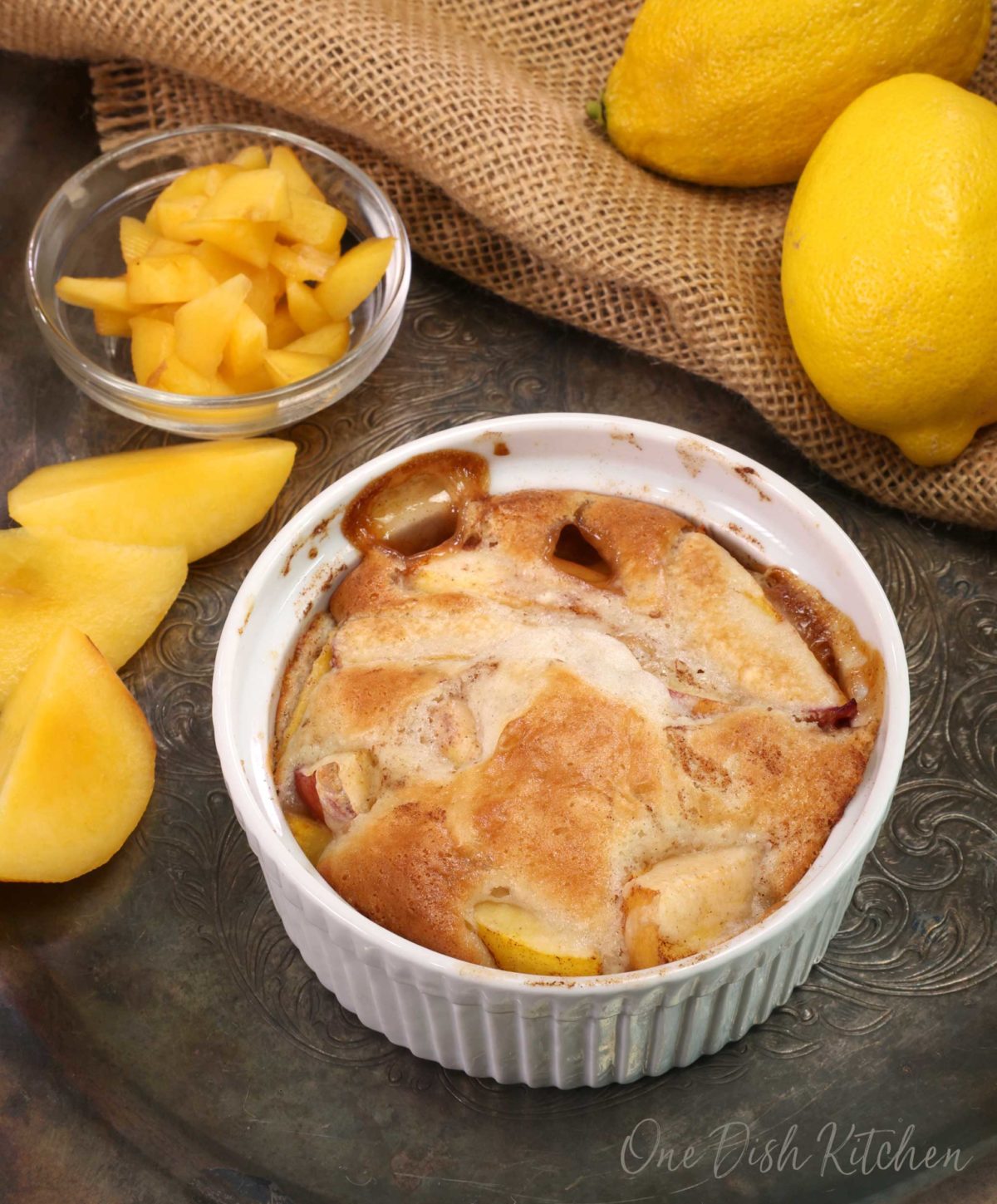 a mini peach cobbler in a small white ramekin on a silver tray next to lemons and fresh peaches.