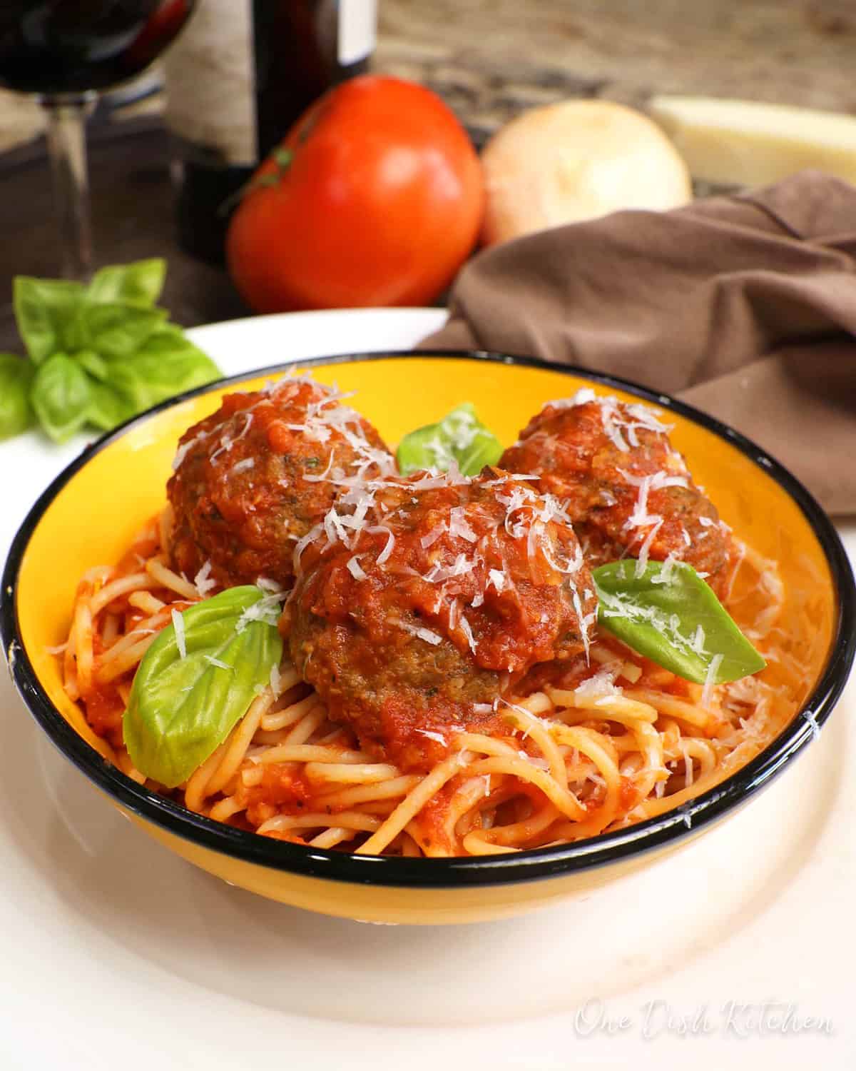 a bowl of spaghetti topped with three meatballs and fresh basil on a white plate next to a bottle of red wine.