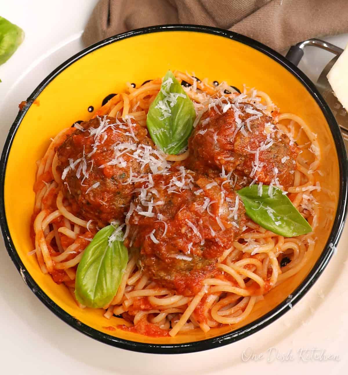 a yellow bowl filled with spaghetti and meatballs next to a wedge of fresh parmesan cheese and a brown napkin.