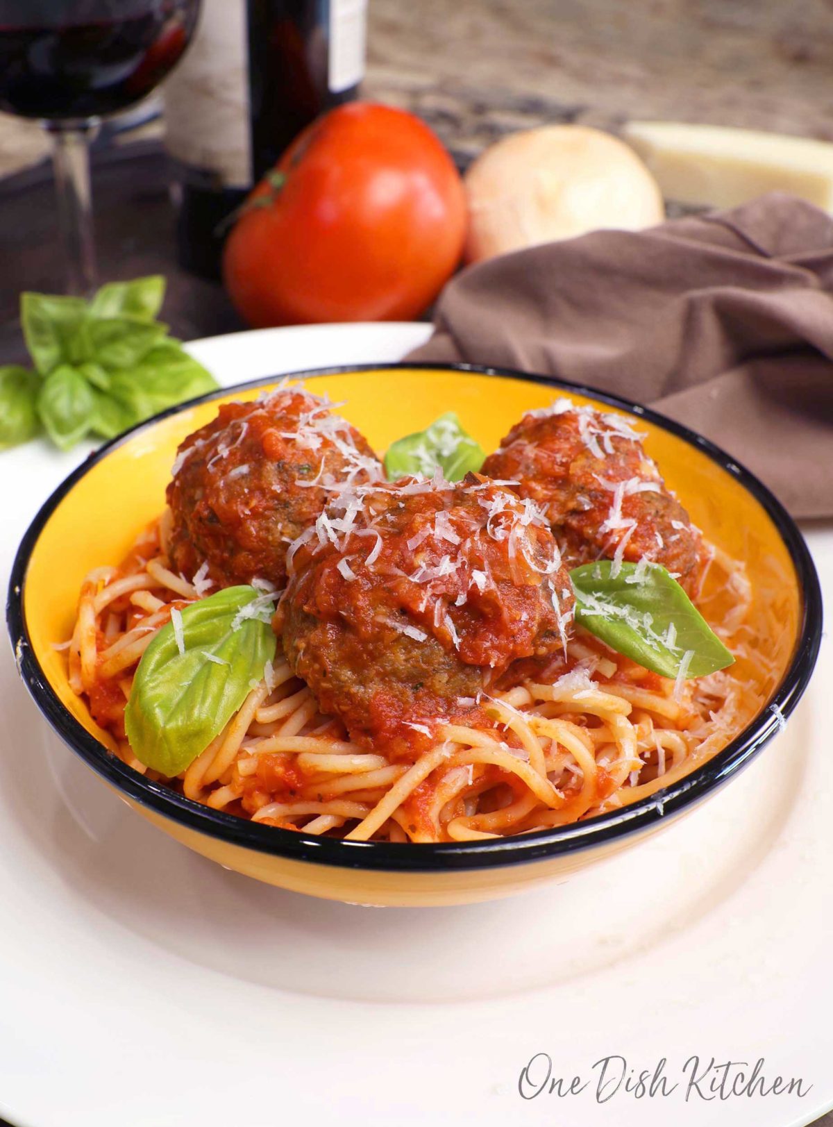 a bowl of spaghetti topped with three meatballs and fresh basil on a white plate next to a bottle of red wine.