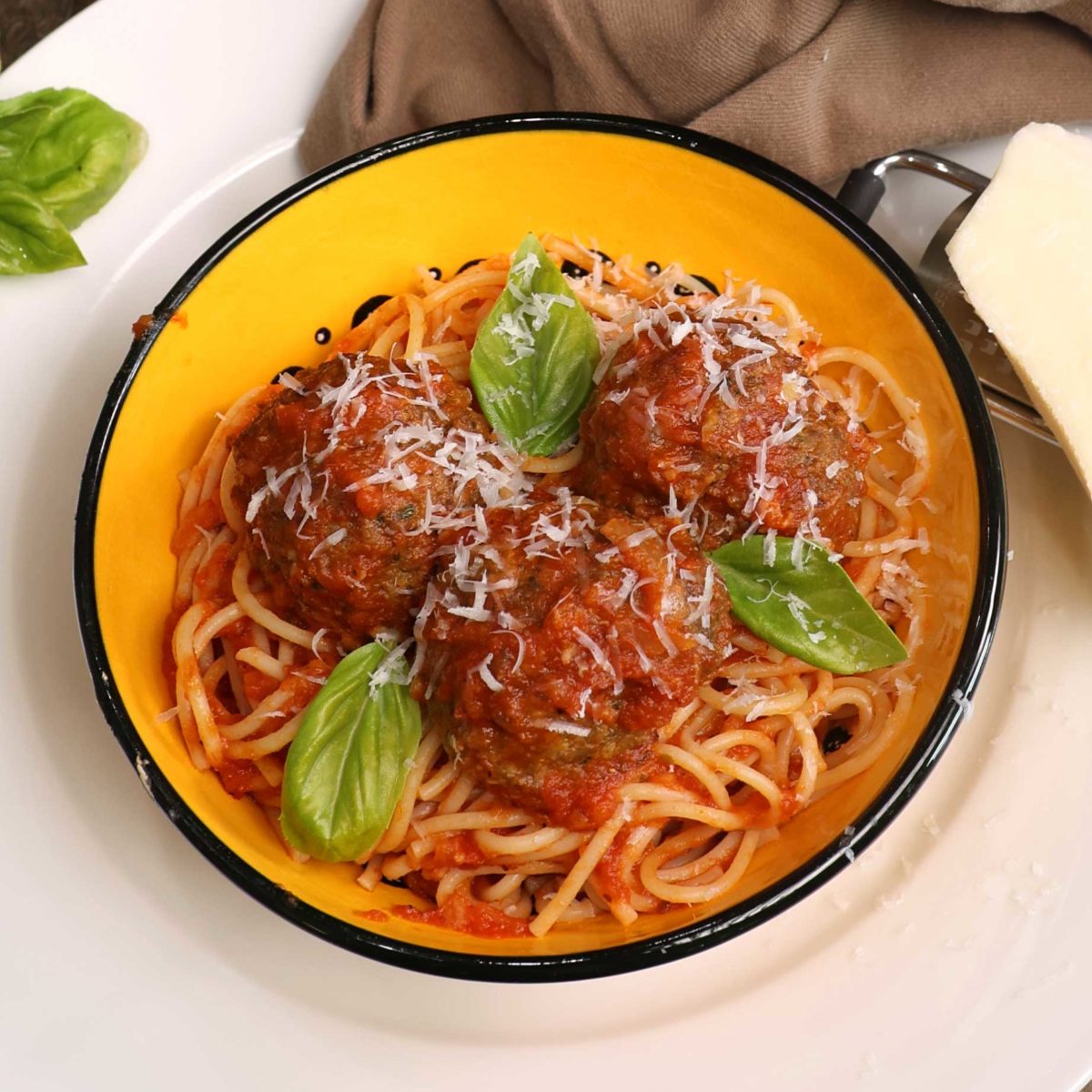 a yellow bowl filled with spaghetti and meatballs next to a wedge of fresh parmesan cheese and a brown napkin.