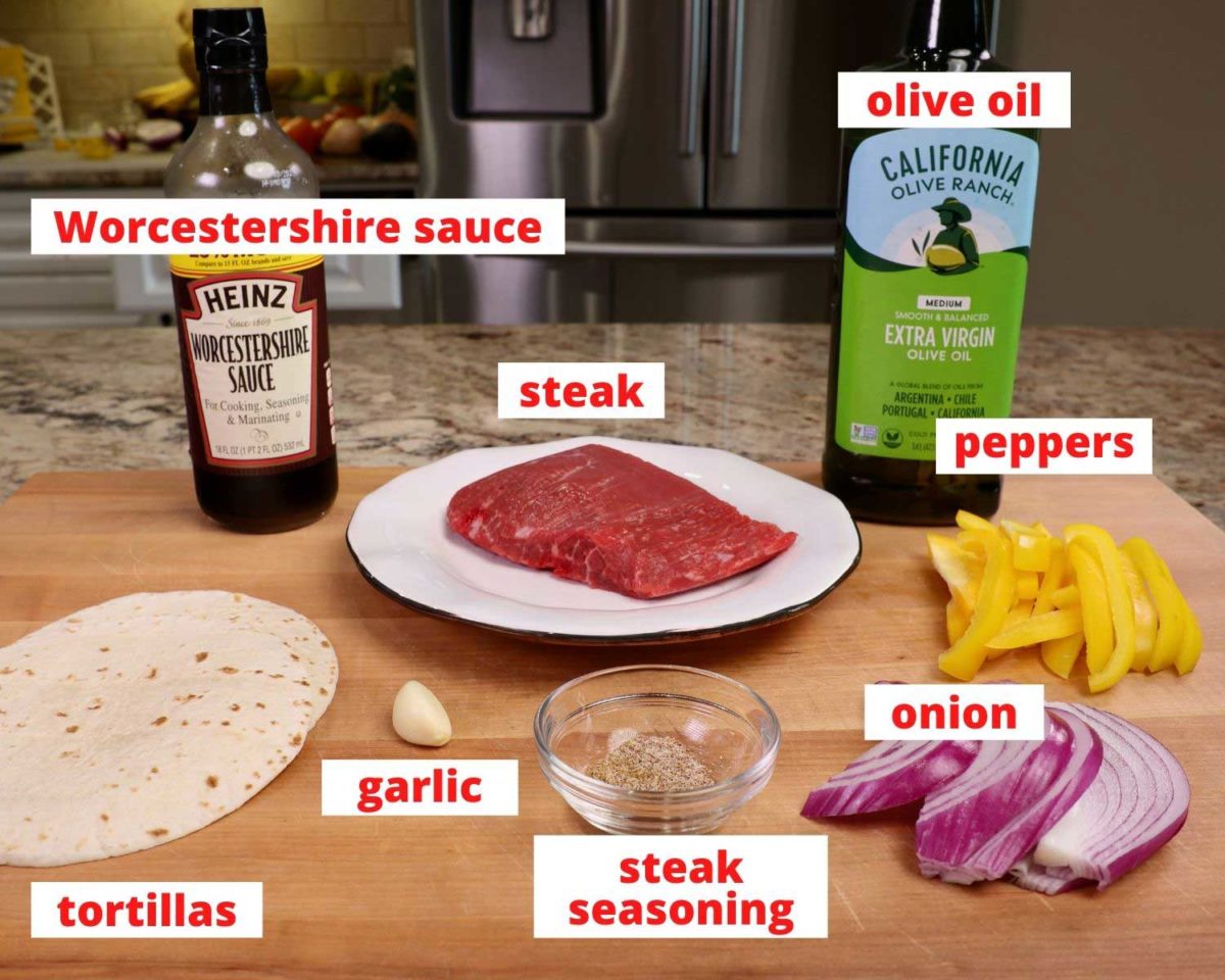 raw steak, worcestershire sauce, tortillas, onions, peppers, and olive oil on a large wooden cutting board.