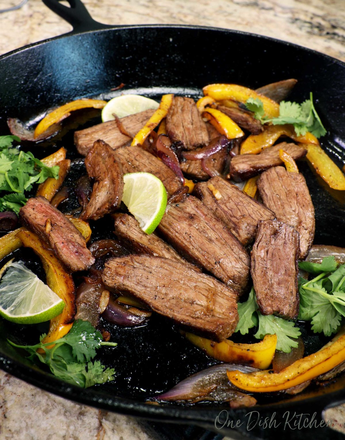 steak fajita meat in a cast iron skillet with vegetables, cilantro, and lime slices scattered around the pan.