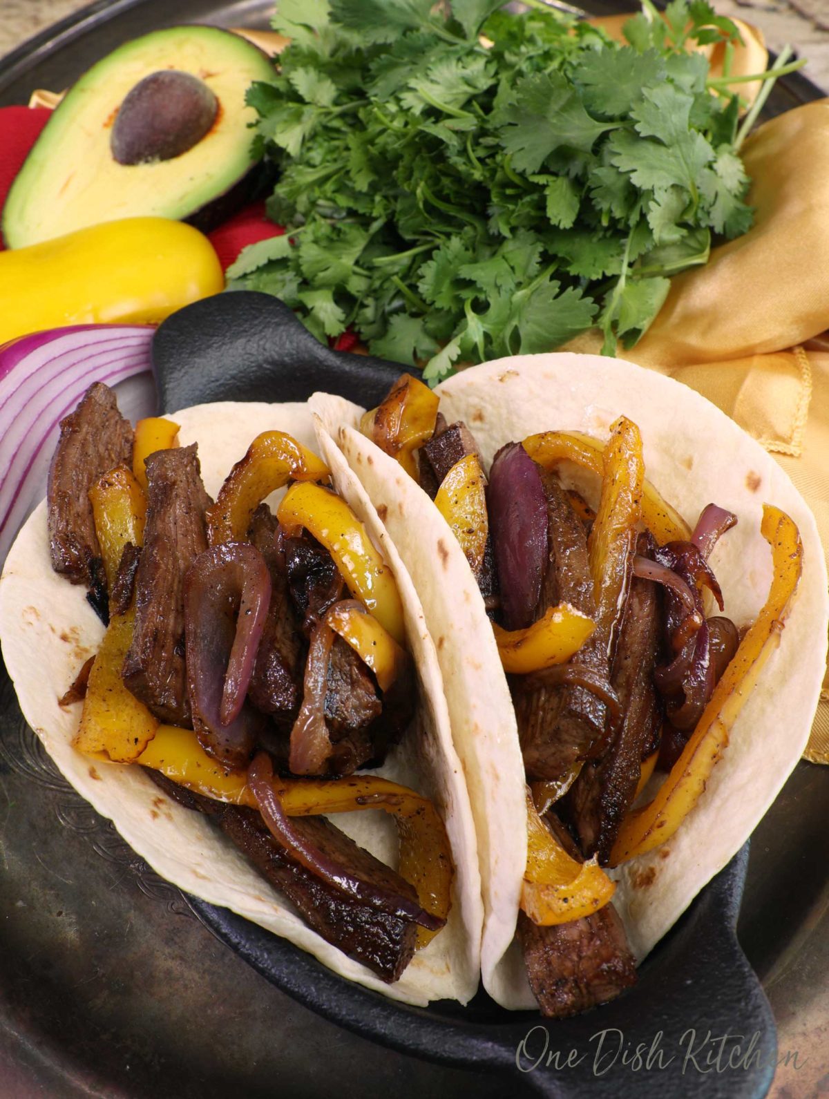 two beef steak fajitas on a black plate next to a half of an avocado and a bunch of cilantro.