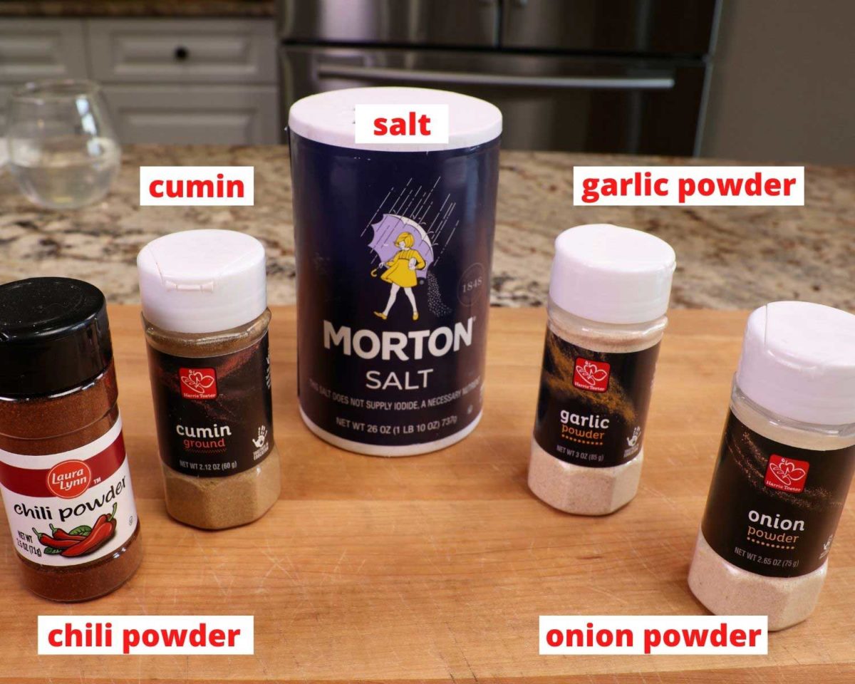 bottles of chili powder, cumin, salt, garlic powder and onion powder on a wooden cutting board in a kitchen.