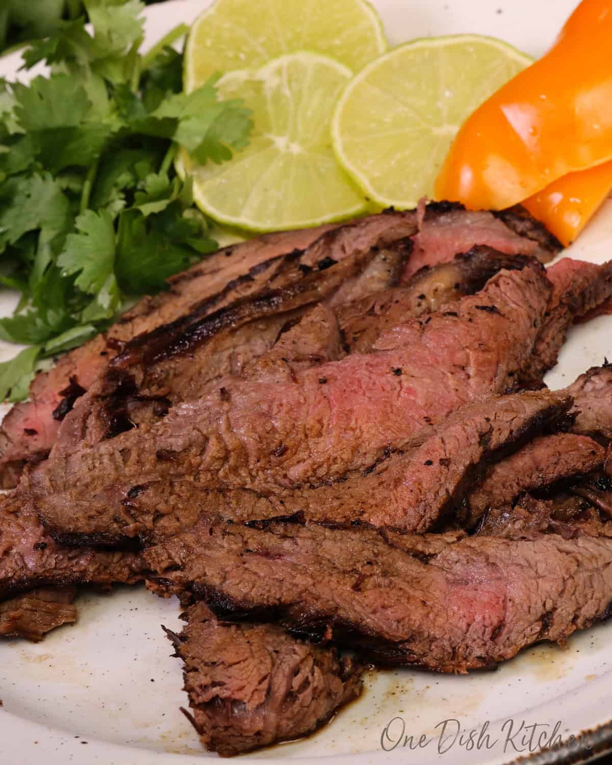 carne asada on a white plate next to cilantro, lime slices, and sliced peppers.