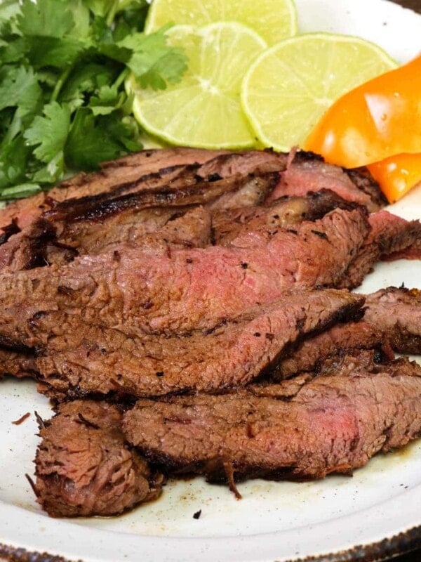 sliced carne asada on a white plate next to cilantro, limes and orange peppers
