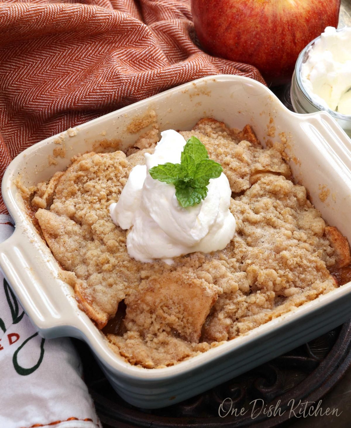 a mini dutch apple pie in a small square blue baking dish next to an orange napkin, a jar of whipped cream and one red apple.