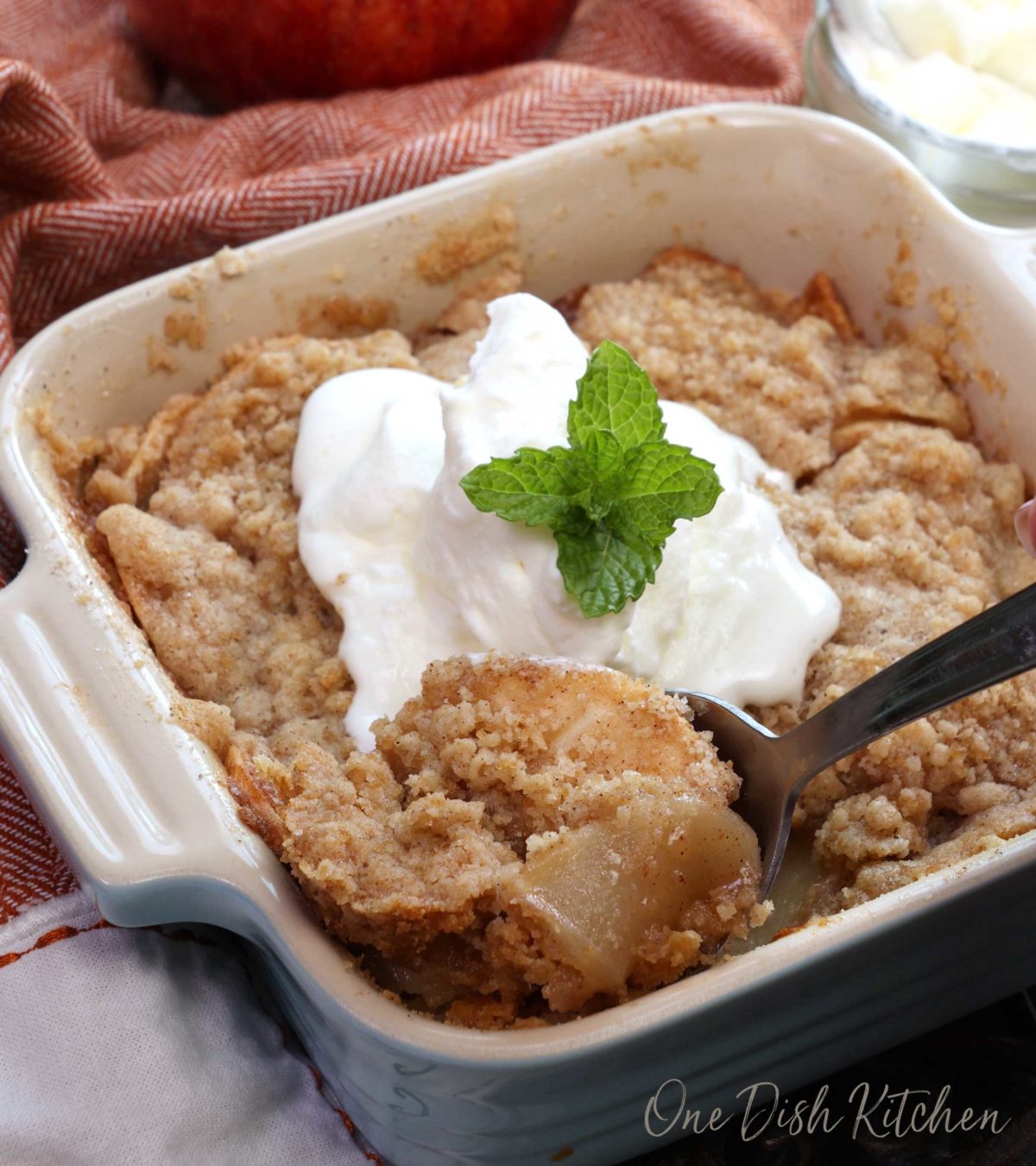 a small dutch apple pie with crumble topping topped with whipped cream and a sprig of mint.