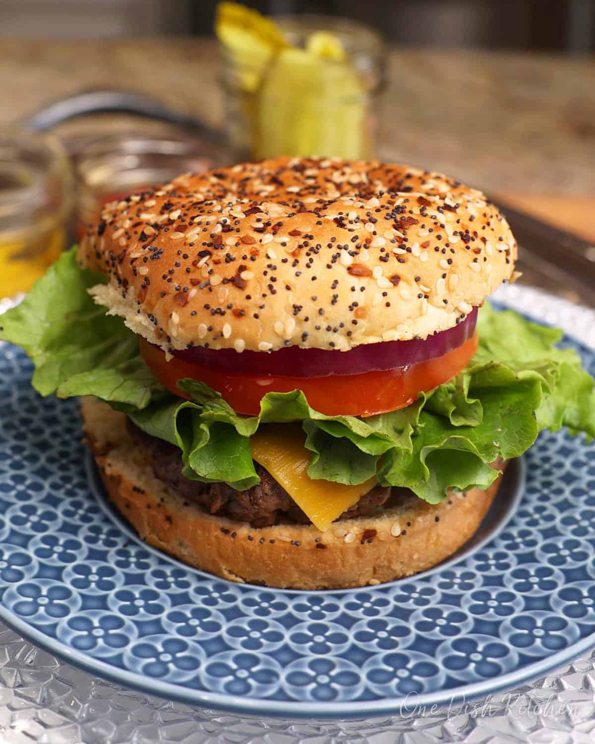 a hamburger on a blue plate next to a jar of pickles.