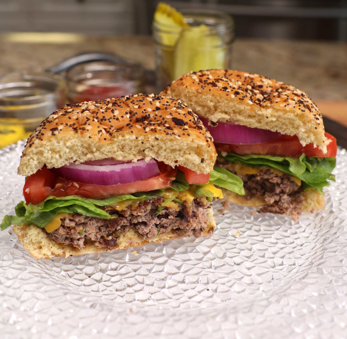 a hamburger topped with cheese, lettuce, tomato, onions and ketchup next to a jar of pickles.