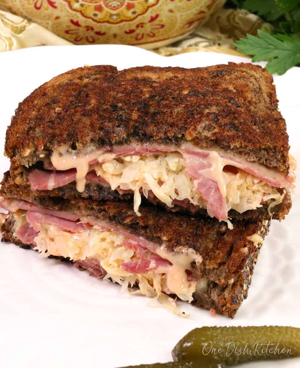 a reuben sandwich on a white plate next to a pickle.