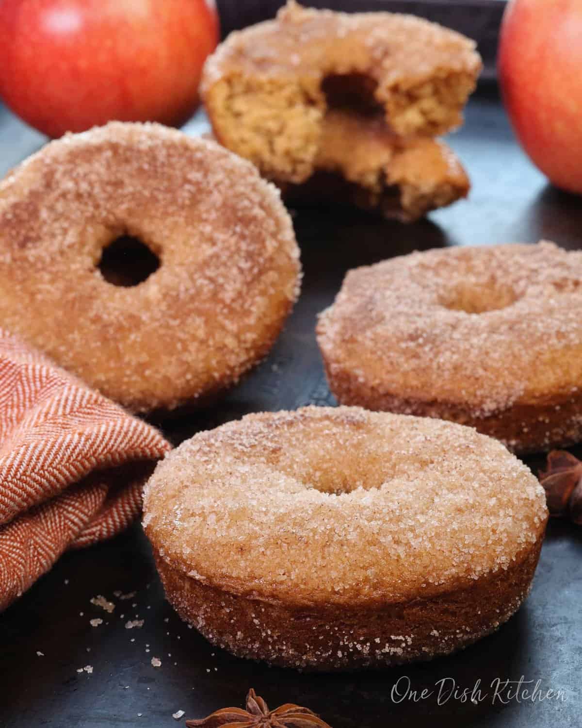 four apple cider donuts with a dusting of cinnamon sugar next to two apples and a star anise.