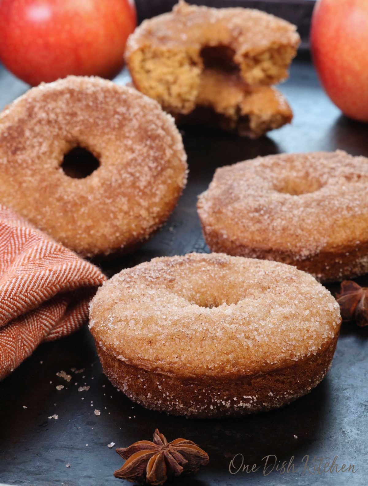 four apple cider donuts with a dusting of cinnamon sugar next to two apples and a star anise.