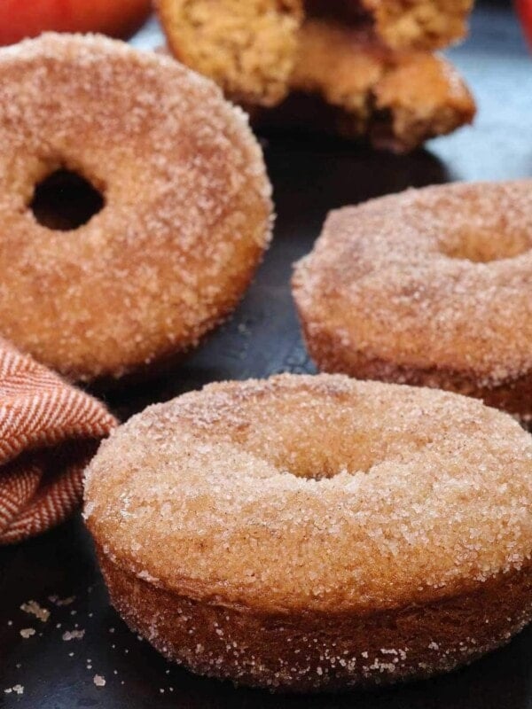 four apple cider donuts with a dusting of cinnamon sugar next to two apples and a star anise.